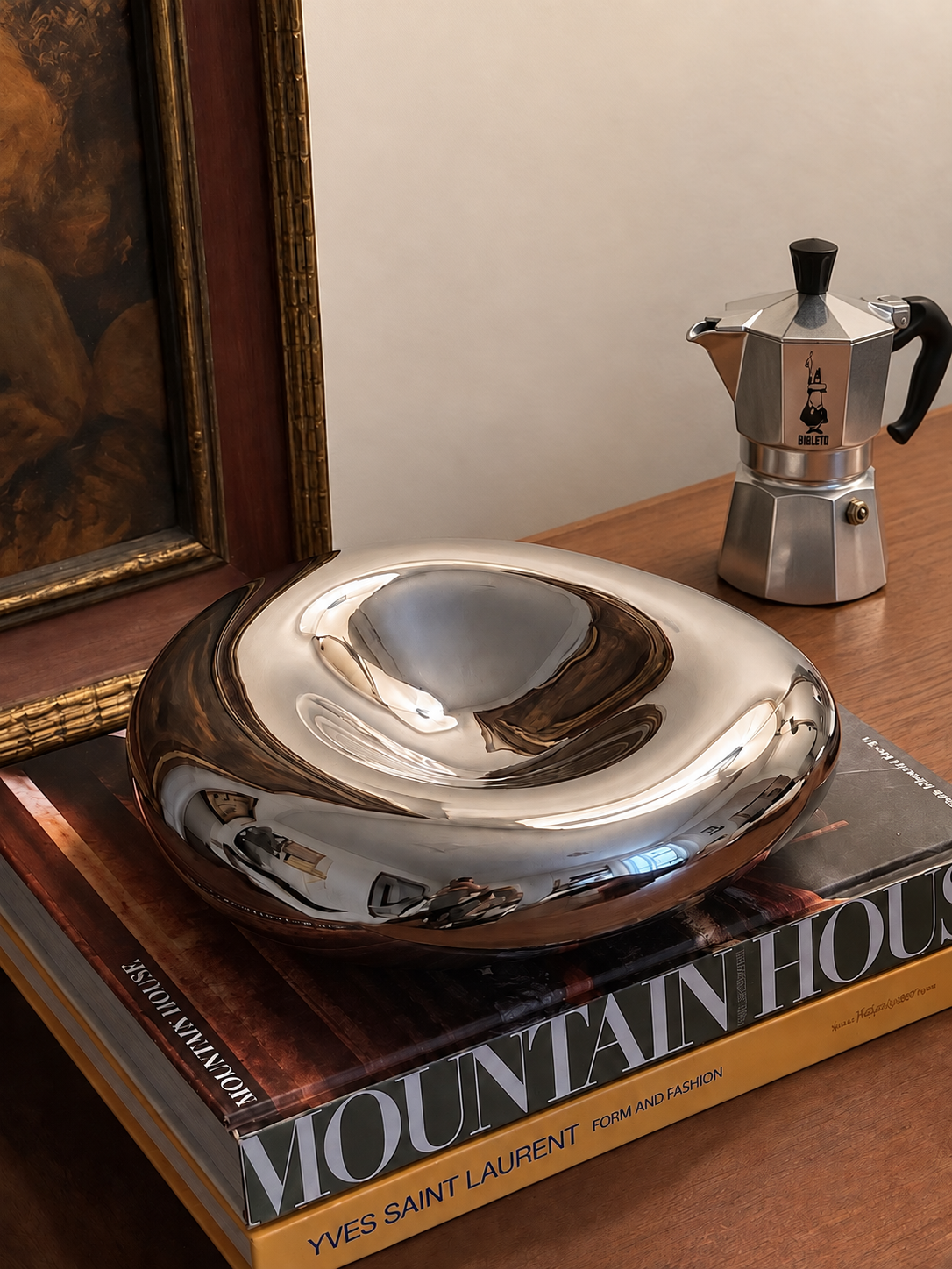 A polished chrome sculptural bowl on an Yves Saint Laurent book, beside a moka pot and a framed painting